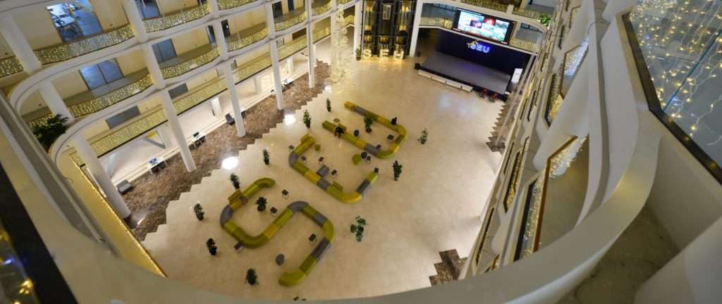 A spacious atrium featuring curved yellow and gray seating, plants, and illuminated railings, with a stage and screens visible at the far end.