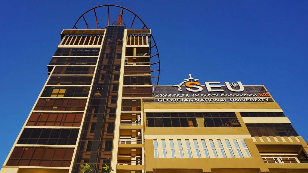 The Georgian National University building features modern architecture with large windows, a distinctive tower, and a clear blue sky backdrop.