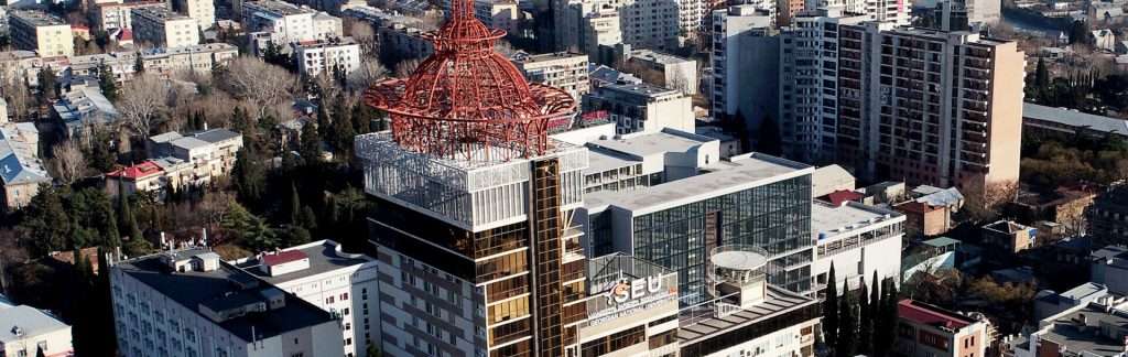 Aerial view of a modern building with a unique red spire, surrounded by urban structures and greenery in a cityscape.